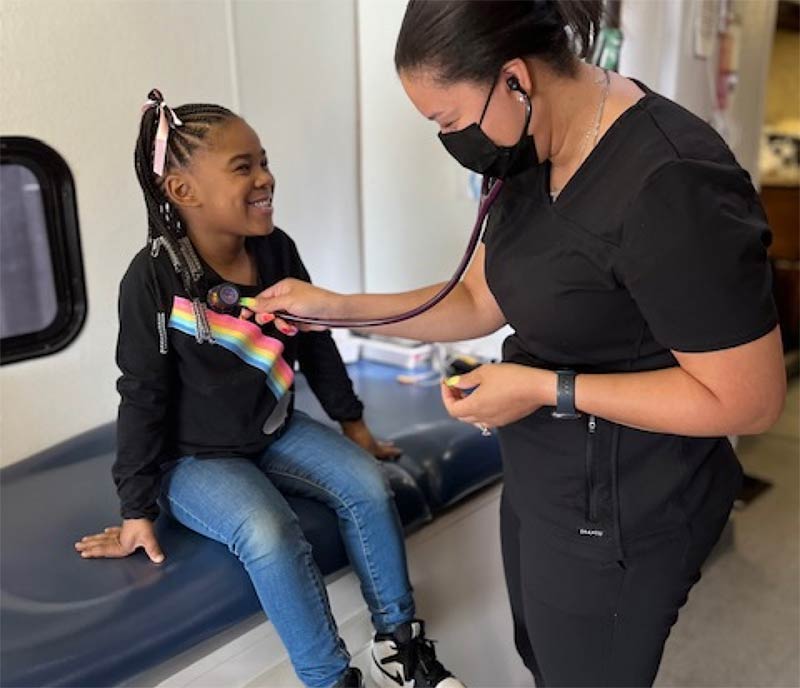 A nurse checks on a smiling girl's heartrate with a stethoscope.